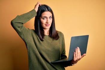 Young brunette woman with blue eyes working using computer laptop over yellow background confuse and wondering about question. Uncertain with doubt, thinking with hand on head. Pensive concept.