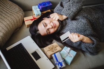 A person laying on a couch holding a credit card next to an open laptop with small wrapped gift boxes around them.