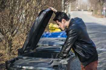 Attractive young man putting out rubbish standing with the lid up on a bin in a row of garbage bins at the side of a street