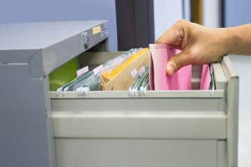 A person's hand is shown reaching into an open file drawer and removing a pink file folder.