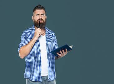 Bearded man who appears to be pondering something is biting on the end of a pen and holding an open notebook.