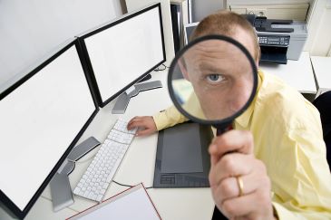 The eye of a businessman sitting behind a dual screen computer seen through a magnifying glass 